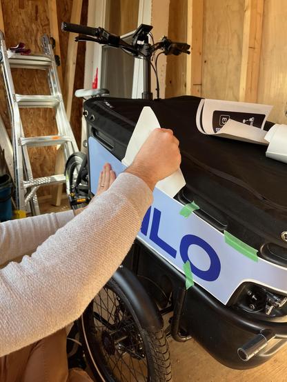 Two hands sticking a decal on the side of a cargo bike box / Deux mains installant un autocollant sur le côté de la boîte d'un vélo-cargo.