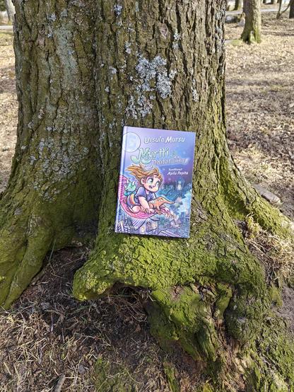 Kuvassa Myrtti ja noitatukka -kirja kuvattuna puuta vasten.