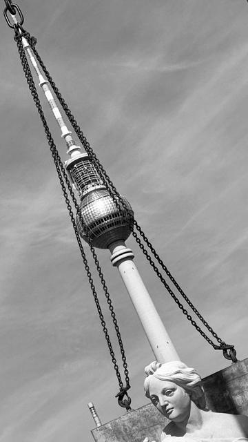A black and white image featuring a prominent television tower partially obscured by chains, with a statue of a woman in the foreground. The background shows a clear sky.