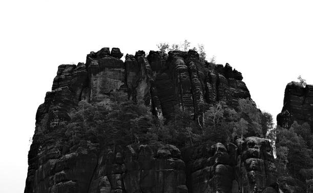 Schwarz-weiß-Fotografie eines Tafelberges aus Sandstein aus der Ferne. Im Vordergrund ist ein Plateau, welches von Bäumen bewachsen ist. Der Stein ist größtenteils fast schwarz, nur fleckenweise kommt ein hellerer Grauton durch. In der Bildmitte ist das obere Plateau des Tafelberges, welches nahezu kahl gegen den fast schneeweißen Himmel im Hintergrund steht.