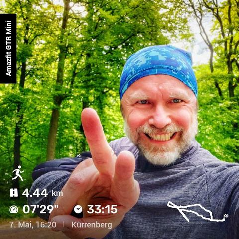 Das Bild zeigt eine Person mit einem blauen gemusterten Stirnband und einem grauen langärmeligen Shirt, die in einem Waldgebiet steht. Die Person hält zwei Finger in einem Peace-Zeichen hoch. Über das Bild ist Text eingeblendet, der Details zu einem Lauf oder Spaziergang zeigt, die mit einer Amazfit GTR Mini Smartwatch aufgezeichnet wurden. Die eingeblendeten Daten sind:  

- ‚Amazfit GTR Mini‘ auf der linken Seite  
- Ein Lauf-Icon mit ‚4,44 km‘  
- Eine Stoppuhr mit ‚07'29"‘  
- Eine Uhr mit ‚33:15‘  
- ‚7. Mai, 16:20 | Kürrenberg‘ am unteren Rand  
- Eine kleine Karte, die die Route des Laufs oder Spaziergangs darstellt.