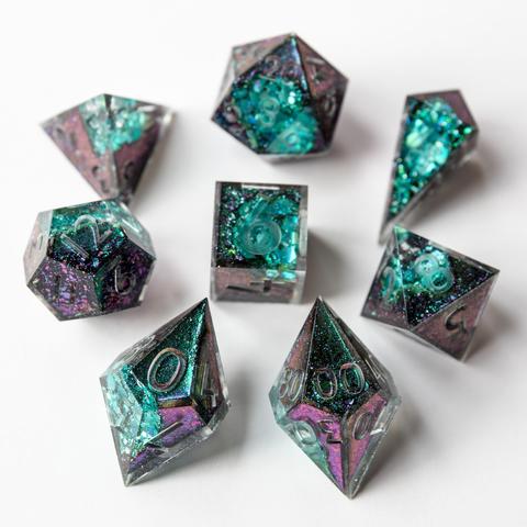 Ein Würfelset in lila/grünem Colorshift mit einer Geode-Seite in Grün. Das Set besteht aus einem D4-Shad, einer D4-Pyramid, D6, D8, D10, D100, D12 und D20. Das Set liegt auf weißem Untergrund. Die Würfel sind im Bild um den D6 angeordnet. Das Set ist noch Work in Progress und noch nicht geschliffen, poliert oder bemalt. Der D10 und der D00 sind im Vordergrund scharf dargestellt. Die anderen Würfel sind im Hintergrund leicht unscharf zu sehen.