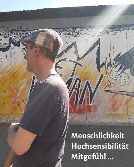 Vor einem Teil der Eastside Gallery, einem Rest der Berliner Mauer mit einem gesprayten Bild, rot-gelbe Flammen unten, oben schwarze Zackenlinien und angedeutete Wolken. Stefan Wetterau steht seitlich vor der Mauer, dunkles T-Shirt und Karo-Mütze, und schaut zur Mauer. Unten rechts die Worte Menschlichkeit, Hochsensibilität, Mitgefühl