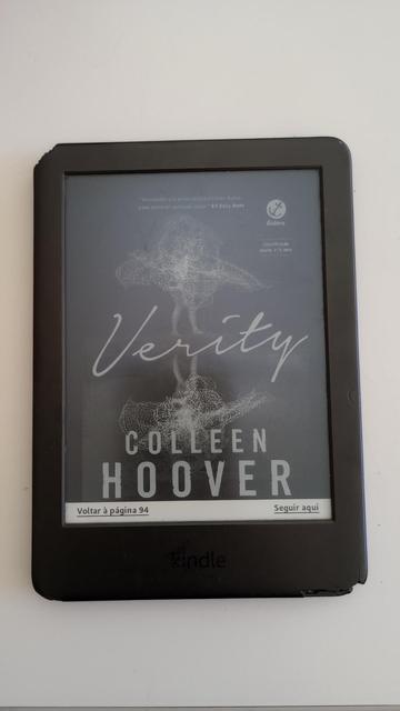 A imagem mostra um Kindle, um dispositivo de leitura eletrônica, com a capa do livro "Verity" de Colleen Hoover. A tela do Kindle exibe a capa do livro, que é predominantemente cinza com um design abstrato em branco no centro. O título "Verity" está escrito em letras grandes e cursivas, enquanto o nome da autora, "Colleen Hoover", está em letras maiúsculas e brancas abaixo do título. No canto superior direito, há um logotipo da editora Galera e uma classificação indicativa de "M+18". No canto inferior esquerdo, há a opção "Voltar à página 94" e no canto inferior direito, "Seguir aqui". A tela também inclui uma citação do "NY Daily News" que diz: "Assim como a jovem em 'Verity', pode ser difícil escapar das suas raízes." O Kindle está posicionado sobre uma superfície branca, destacando o dispositivo e a capa do livro.