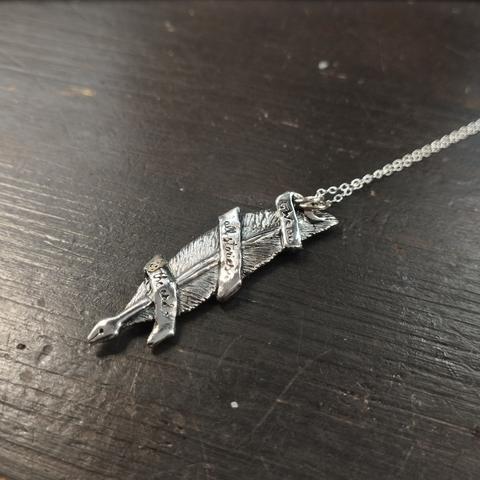Pieza de plata en forma de pluma con plumín para escritura rodeada por una banda donde se lee en tres partes "we are all stories in the end".