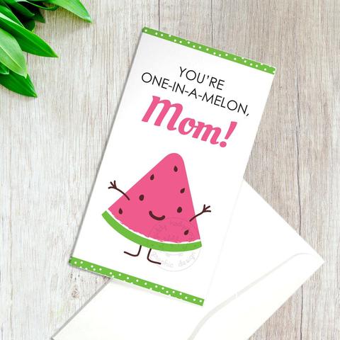 Picture of a greeting card with a smiling cartoon watermelon with a face and arms and legs. The text above says "You're One-In-A-Melon, Mom!" There is a green polka dot border on the top and bottom, and below the card is an envelope. Next to the envelope and card is a red pencil to the bottom right, and to the top left are some leaves.