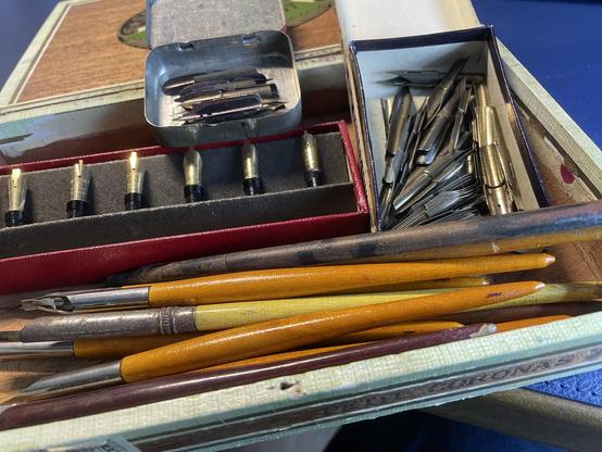 An assortment of vintage writing instruments, including various nibs, fountain pen tips, and wooden dip pens, housed in an open cigar box. The contents show signs of use and age.