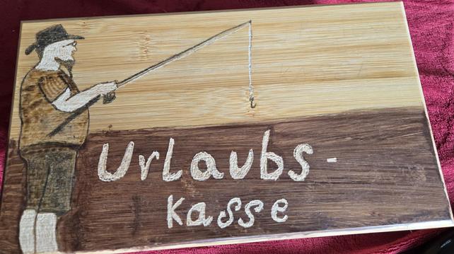 Das Bild zeigt eine Holzkiste, auf der eine Zeichnung eines Anglers mit Angelrute zu sehen ist.  Unterhalb der Zeichnung ist hell "Urlaubs-Kasse" graviert.  Das Brett scheint aus zwei verschiedenen Holzarten zu bestehen, einem helleren oben und einem dunkleren unten. Die Zeichnung und die Beschriftung wirken handgemacht, möglicherweise durch Brandmalerei oder eine ähnliche Technik.