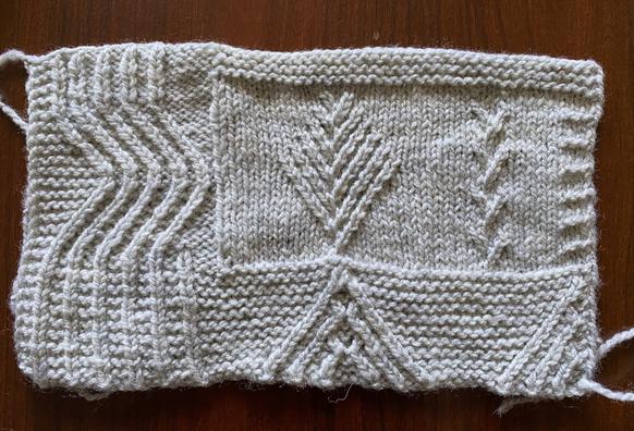 Hand knit swatch in a light bluish grey, with twisted stitch patterns of waves on the left, mountains against garter stitch on the bottom, sort of conifer trees in the middle, and something that when I play around with the placement will hopefully evoke grasslands. The swatch is now blocked, so it lies nice and flat and the stitches are more even.