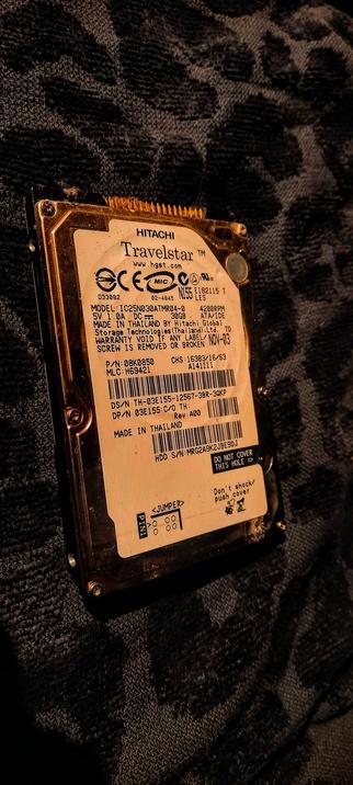 The photograph shows a Hitachi Travelstar hard disk drive (HDD) placed on a dark fabric with a leopard print pattern. The HDD is oriented vertically, with the label facing the viewer. The label is predominantly white with black text and includes various details such as the model number (IC25N030ATMR04-0), capacity (30GB), speed (4200RPM), and manufacturing information (Made in Thailand by Hitachi Global Storage Technologies (Thailand) Ltd.). The label also features certification marks like CE, FCC, and MIC, along with a serial number (D33082) and a barcode. The HDD has a gold-colored connector and is secured with screws. The bottom of the label includes a warning not to cover a specific hole and a caution against shocking or pushing the cover. The overall condition of the HDD appears to be good, with no visible damage.

 Ovis2-8B

🌱 Energy used: 0.254 Wh