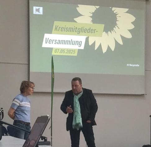 Sonnenblumenlogo auf einer Leinwand "Kreismitgliederversammlung", davor zwei Mitglieder im Zwiegespräch