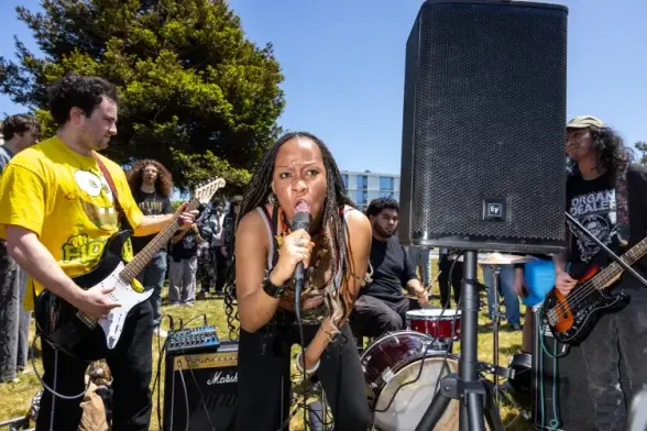 Person screams into mic as band plays outside with crowd watching.