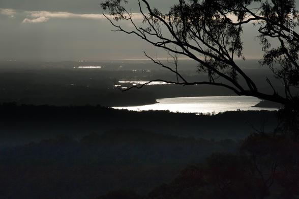 A high view over a dark, wooded plain. Trees and features are barely visible in almost total gloom, but sunlight filtering through heavy cloud shines off ponds and dams like drops of molten silver splashed over the land. A nearby eucalypt frames the top right of the image in silhouette.