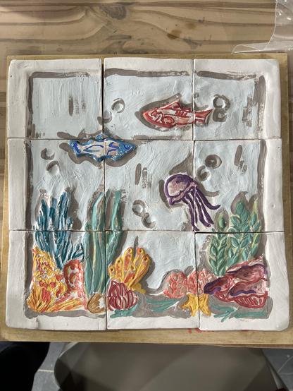 Fotografía de un mosaico en cerámica con detalles del mar. El mosaico tiene nueve cuadrados y dibujados dos peces y un pulpo más el fondo marino de algas, corles y piedras.
