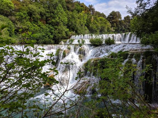 A scenic view of a cascading waterfall in Krka National Park. The water rushes over multiple tiers, creating a beautiful display of white water against a backdrop of lush green trees. The foreground includes some foliage, framing the waterfall. The sky is partly cloudy.