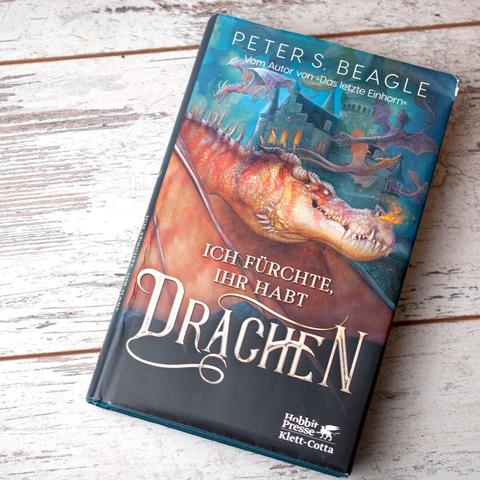 Buch "ich fürchte, ihr habt Drachen" von Peter S. Beagle auf weißem Holzuntergrund, fotografiert von oben.