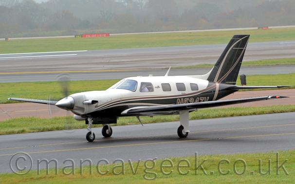 Side view of a white and black, single-engined Bizprop taxiing from right to left along a black taxiway.
The plane has a white upper fuselage with black lower fuselage and tail.
There are light and dark brown stripes running along the fuselage before sweeping up in to the tail.
The brown registration "N5347V" is on the lower rear fuselage.
Strips of green grass separate taxiways from the black runway in the background, with more grass leading up to trees vanishing in to haze in the distance.