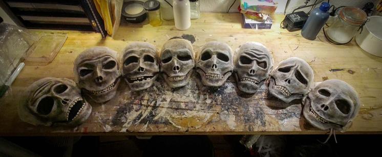 This image shows a row of eight handcrafted skull masks or decorations arranged on a wooden workbench. The skulls appear to be made of paper maché and have been painted a weathered grayish-white color. Each skull has distinctive features and expressions - some with teeth showing, others with different facial structures and contours. The workbench has paint stains and signs of artistic work, with various containers, tools, and supplies visible in the background, suggesting this is in an artist's workshop or craft space. This appears to be a Halloween, Day of the Dead, or theatrical prop-making project in progress.