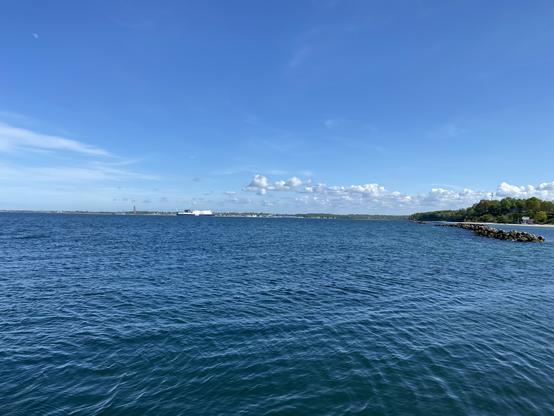 Leicht gewellte Kieler Außenförde bei strahlendem Sonnenschein, aufgenommen vom Fähranleger Schilksee. Rechts der Strand von Schilksee mit vorgelagertem Wellenbrecher. Mittig eine DFDS-Fähre auf dem Weg in den Hafen. Dahinter Laboe mit dem Marine-Ehrenmal. Über dem Kreis Plön liegt parallel zum Horizont rechts vom Ehrenmal eine Reihe an knuffigen Quellwolken.