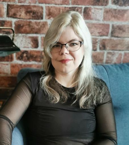 White woman with white-blonde hair just past her shoulders, a black top, and glasses, sitting on a teal sofa with a red brick wall behind her.