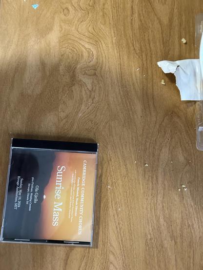 The image depicts a compact disc (CD) placed on a wooden surface. The CD appears to be a music album titled "Sunrise Mass," featuring the Cambridge Community Chorus. The CD case is transparent with a black border, displaying the album cover, which shows a sunrise or sunset scene with warm, orange hues. The surrounding area includes a few scattered crumbs or small particles on the wooden surface, suggesting a casual or informal setting.