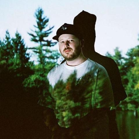 A white man in a black ballcap and a white t-shirt stands against the wall with an image of a forest projected onto him. He has stubble and looks directly into the camera.