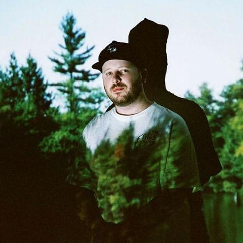 A white man in a black ballcap and a white t-shirt stands against the wall with an image of a forest projected onto him. He has stubble and looks directly into the camera.