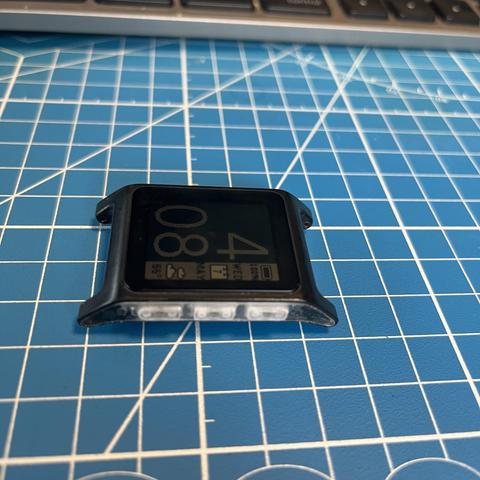 The image shows a black smartwatch with a square face, placed on a blue cutting mat with a white grid pattern. The smartwatch displays the time "4:08" in large digits on its screen, along with additional information such as the date and possibly other icons or notifications. The watch has a black casing with a slightly curved bottom edge, and the brand name "Pebble" is visible on the lower part of the watch face. The background includes a partial view of a keyboard, suggesting the setting might be a workspace or desk area. The smartwatch is positioned centrally on the mat, with the grid lines providing a structured backdrop.