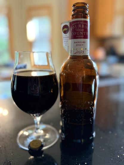 A tall snifter glass full of dark beer next to a tall brown bottle with a white neck label. A deep purple section has the nom de plume and a small flag has the goose logo