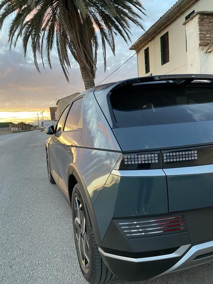Un coche Hyundai ioniq 5 y eléctrico está aparcado en una carretera con una palmera cerca. El fondo presenta un cielo al atardecer con colores vibrantes. Se puede ver un edificio en la distancia, lo que se suma a la atmósfera serena.