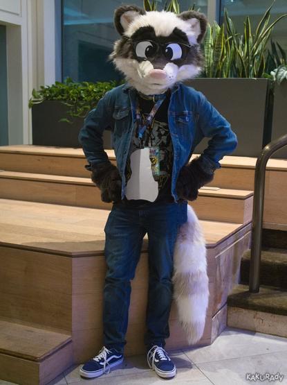 A tan and dark brown toony raccoon fursuiter wearing black glasses, demin shirt with a printed black T-shirt, jeans, and black skateboarding shoes with white soles, in front of planters on waist-height wooden steps leading up to a glass wall beside stairs