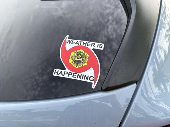 A blue hatchback with a Weather is Happening sticker on the corner of the rear window. The sticker is a bright red hurricane symbol with a yellow hexagon and an Illuminati-like pyramid drawing in the center.