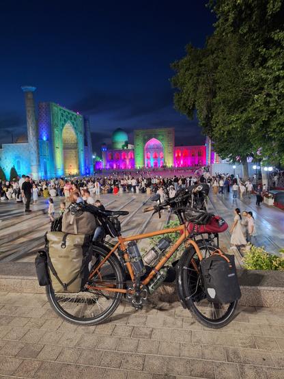 Bepacktes Fahrrad auf dem Registanplatz in Samarkand, im Abenddunkel. Die drei Medresen sind grellbunt beleuchtet: in Pink und blau vor allem.