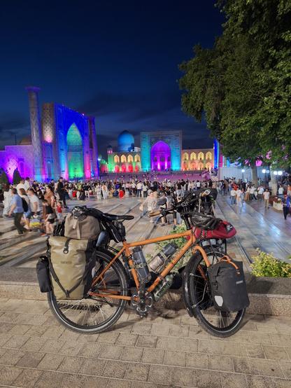 Bepacktes Fahrrad auf dem Registanplatz in Samarkand, im Abenddunkel. Die drei Medresen sind grellbunt beleuchtet: in türkis, gelb und pink vor allem.