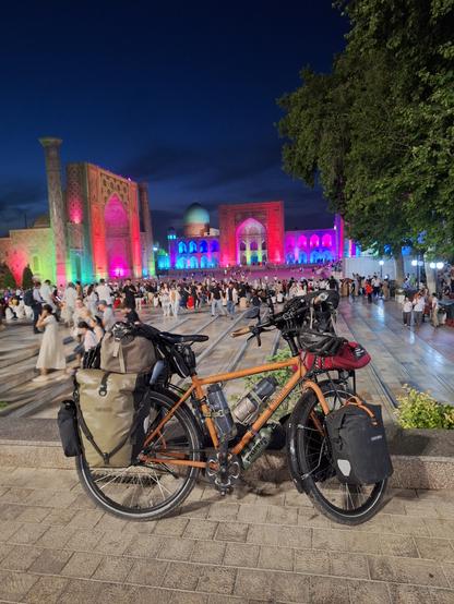 Bepacktes Fahrrad auf dem Registanplatz in Samarkand, im Abenddunkel. Die drei Medresen sind grellbunt beleuchtet: in Orange, rot und pink vor allem.