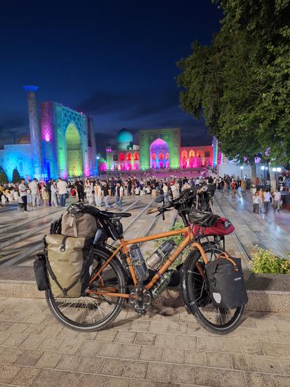 Bepacktes Fahrrad auf dem Registanplatz in Samarkand, im Abenddunkel. Die drei Medresen sind grellbunt beleuchtet: in grün, blau und pink vor allem.