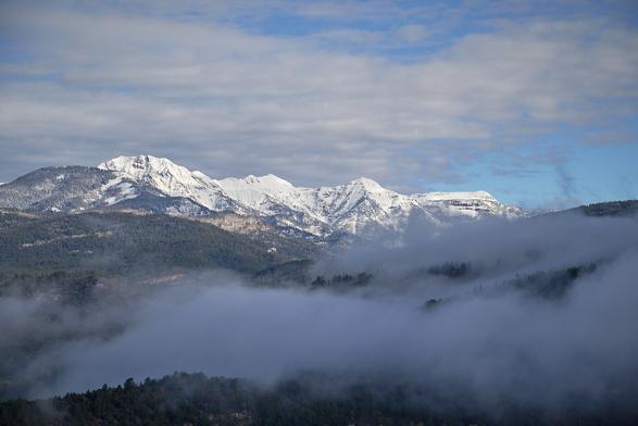 Snow-capped mountains under a partly cloudy sky, with mist drifting over the forested hills below, creating a peaceful and majestic spring-in-the-mountains scene.