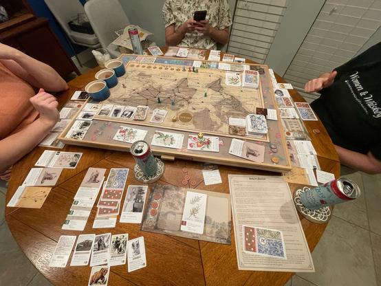 The photo shows a round table with three people around it, and on the table is a board game with a bunch of cards and game pieces.