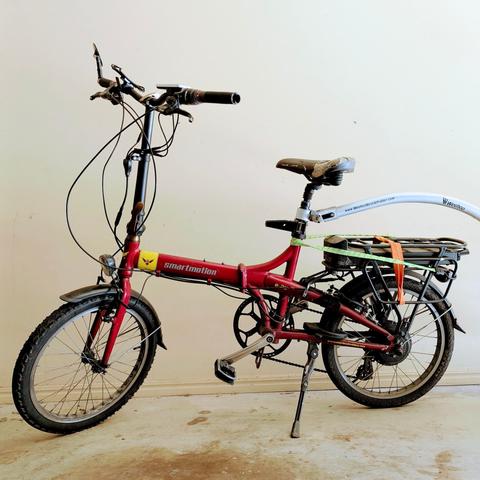 A red Smartmotion E20 folding e-bike is parked against a light-colored wall. The bike has a black seat, handlebars, and a rear rack with an orange and green strap securing something. A white, curved bar with text on it extends from the rear of the bike.