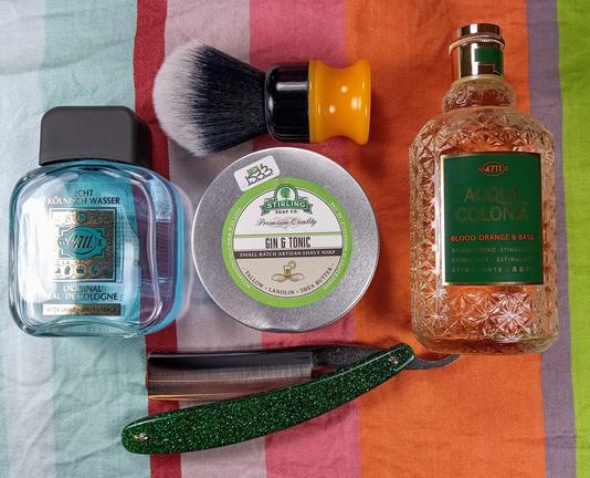 (Le Chat Mistral description)

The image showcases a collection of grooming items arranged on a striped cloth. On the left, there's a transparent bottle of "Echt Kölnisch Wasser" cologne. In the centre, a tin of "Gin & Tonic" shaving soap from Stirling Soap Co. is visible, accompanied by a shaving brush with a black and yellow handle. Below the soap, there's a straight razor with a green, glittery handle. On the right, a bottle of "Acqua di Colonia" cologne with a textured, amber-coloured liquid is displayed. The items are neatly organised, suggesting a traditional shaving setup.