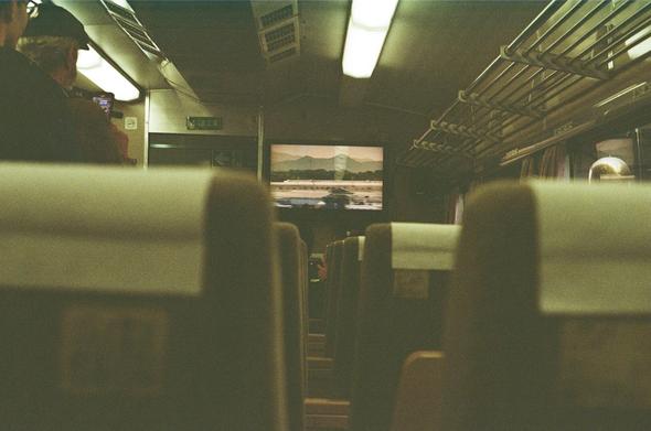 This image depicts the interior of a train carriage. The perspective is from the back of the carriage, looking forward. The seats are arranged in rows, with a central aisle leading towards the front. Overhead, there are luggage racks and fluorescent lighting. A screen at the front displays a scenic view, possibly of a landscape with mountains and water, suggesting a travel destination or route. The image is slightly blurred, giving a sense of motion or speed. The overall ambiance is that of a modern, comfortable train journey. This is the bullet train reproduction is at the York National Railway Museum.