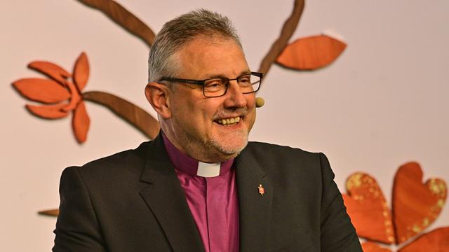 Ein Mann mit graumeliertem Haar, Brille, Kollarhemd, dunklem Jackett und Bischofsnadel am Revers schaut freundlich lächelnd, vielleicht erheitert, in die Zuhörerschaft.