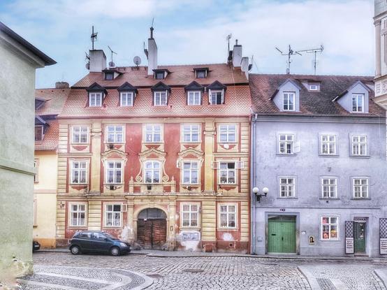 Farbige Bürgerhäuser, links rot, gelb, rechts hellblau, in der tschechischen Stadt Cheb. Der blaue Himmel darüber nur leicht bewölkt.