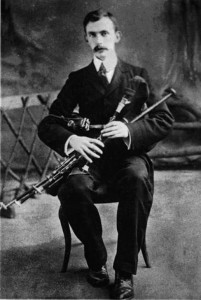 Éamonn Ceannt sitting on a chair holding a set of uilleann pipes.