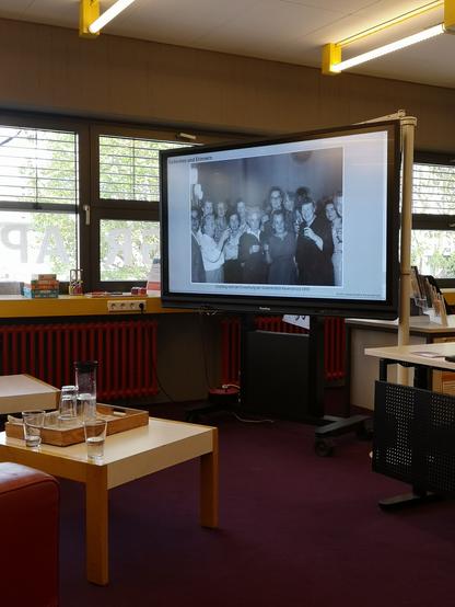Foto auf dem Monitor von der Einweihung der Gedenkstätte Ravensbrück, viele Frauen und wenig Männer prosten der Kamera zu, darüber steht Gedenken und Erinnern.
Zu sehen ist noch ein Teil der Sitzecke in der Bibliothek, ein Tisch mit Gläsern und Wasser.