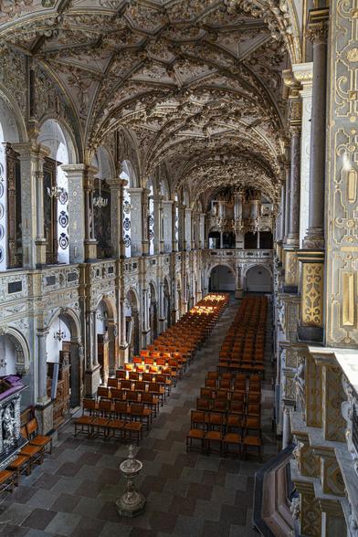 We're on the upper balcony level that surrounds an ornately decorated medieval chapel. It's a large space, rectangular in form, and we're looking from one of the front corners diagonally across and down towards the opposite rear corner. The flooring is tiled with large squares of grey and dusky pink but it's not a chequerboard effect, instead having each row of alternating colours just offset a little from the one before it so that the tiles appear to create jagged lines across the space. There are two columns of chairs set out in rows either side of a narrow aisle. Each column has around thirty rows of four, orange-padded seats. A small silver font or statue is below us, in front of the aisle. The chapel is very tall and this balcony level is about five metres up with arched openings between square, marble columns looking down below. Another five metres reaches the ceiling which is curved across its width down the length of the chapel. Every surface is highly decorated in artwork or gold trim.