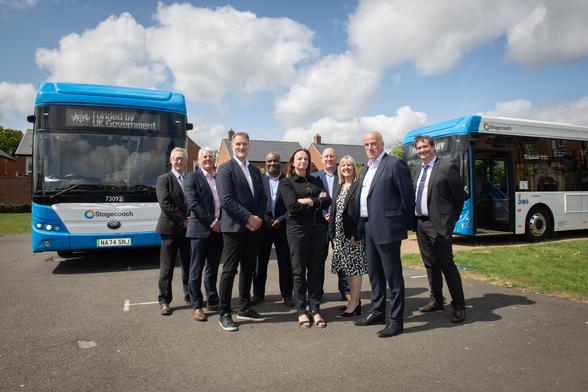 The image shows a group of eleven people standing in a car park, positioned in front of two blue and white buses. The bus on the left features a sign stating it is "Funded by UK Government" and displays the Stagecoach logo along with the number 73092. The bus on the right also bears the Stagecoach logo and other visible numerical details. The individuals, a mix of men and women, are dressed in business attire, and the atmosphere appears formal yet friendly. The sky above is partly cloudy, providing bright and even lighting across the scene.

Photo by Stagecoach