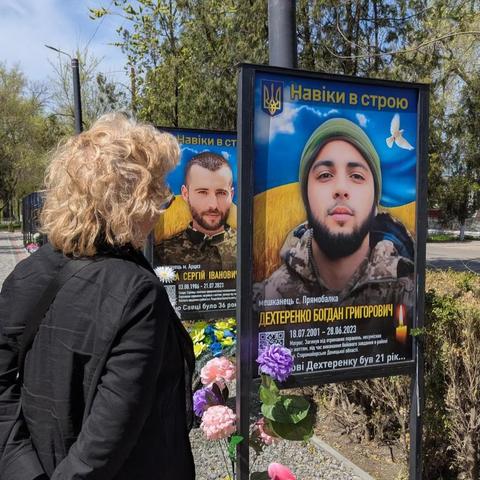 Beate Müller-Gemmeke steht vor Gedenktafeln mit Porträts gefallener Soldaten. Sie blickt auf das Bild eines jungen Mannes, umgeben von Blumen und Fahnen in Blau und Gelb.