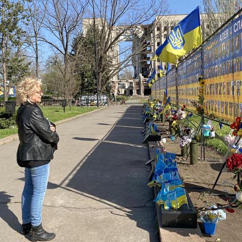 Beate Müller-Gemmeke steht in Mykolajiw vor einer langen Gedenkwand mit Fotos gefallener Soldaten. Dahinter ragt ein von Bomben zerstörtes Gebäude auf. Ukrainische Fahnen wehen im Wind.
