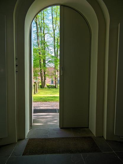 I am standing inside a church. The walls and doorway are light-coloured, the inside of the room is dark. The double-leaf door is curved at the top. The left half of the door is open. The view falls on lawns and green trees in the sunshine. A house made of red bricks can also be seen.